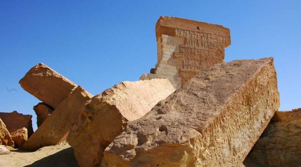 Ancient carved reliefs on the top structure of Amun Temple in Siwa Oasis, juxtaposed against fallen sandstone blocks under a clear blue sky.