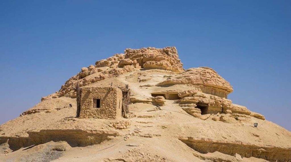 A stone structure with a small building and openings on a rugged hilltop at Gebel al-Mawta in Siwa Oasis, under a clear blue sky.