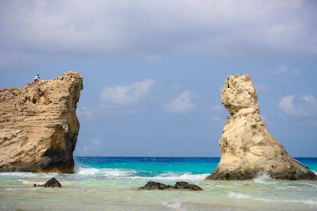 A person sitting atop a rugged limestone rock formation at Cleopatra's Beach, Marsa Matrouh, with turquoise waves crashing against the shore.