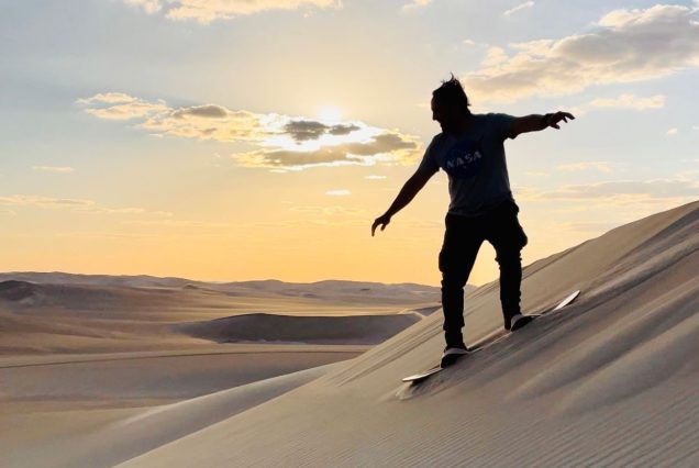 A man balancing on a sandboard while sliding down a dune during sunset, set against the golden sands and soft glow of the sky.