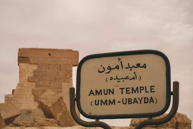 A weathered sign for the Amun Temple (Umm-Ubayda) stands in front of the ancient ruins, with crumbling stone structures in the background.