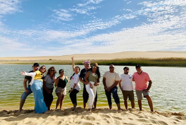 A joyful group of friends standing by a desert oasis, surrounded by calm waters and golden sand dunes under a bright blue sky.