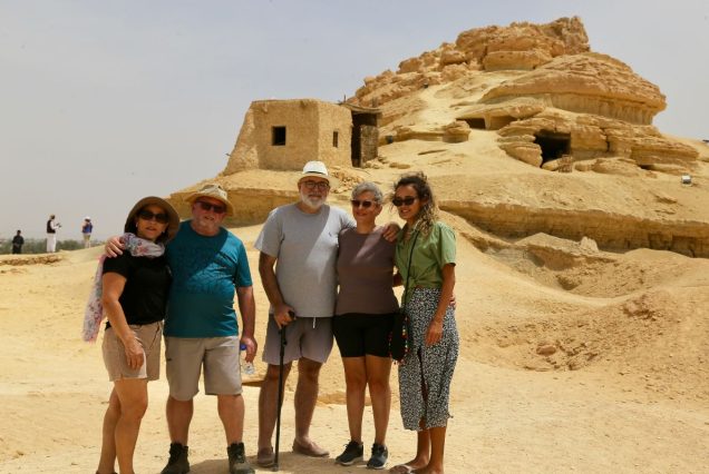 A group of five travelers stands together smiling in front of ancient ruins in the desert, with a sandy hill and a historic structure in the background.