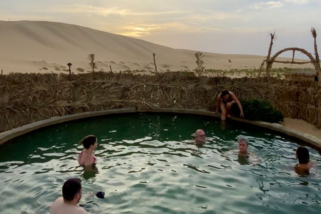 People enjoying a natural desert hot spring pool with sand dunes in the background