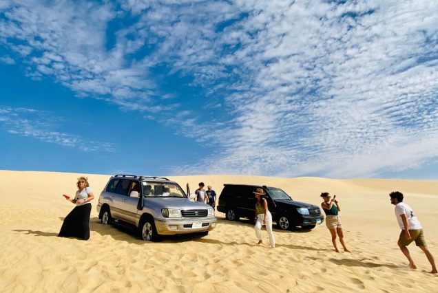 A group of travelers beside two off-road vehicles on a vast desert landscape, under a blue sky scattered with clouds.