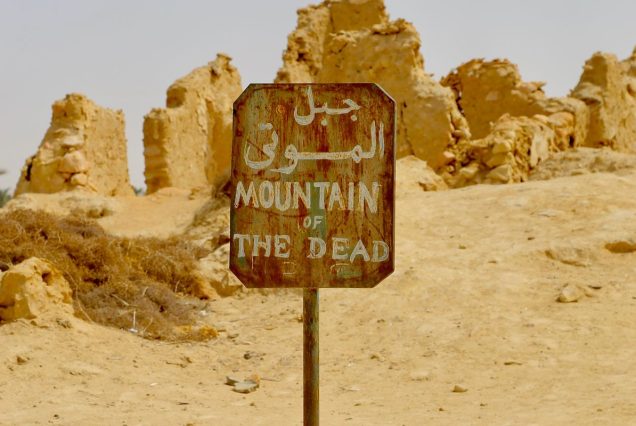 A rusty sign marking the “Mountain of the Dead” stands amidst desert ruins, with weathered stone structures scattered in the background.