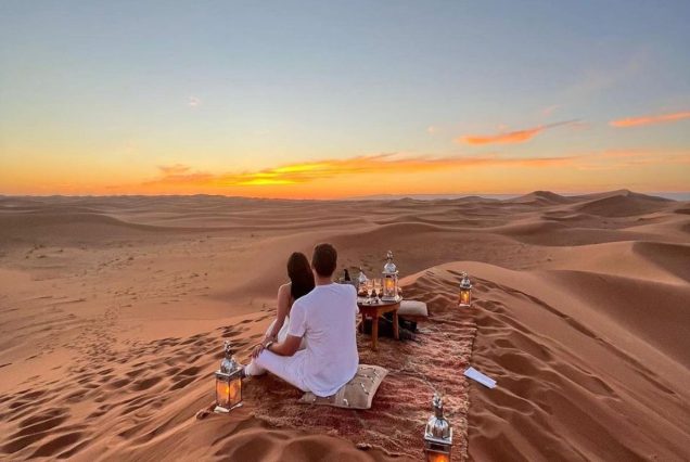 A couple sitting on a blanket atop a sand dune, surrounded by lanterns, watching the sunset over the desert.