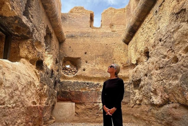 A tourist dressed in black gazes up while standing inside an ancient Egyptian stone structure, with the walls showing signs of weathering and decay.