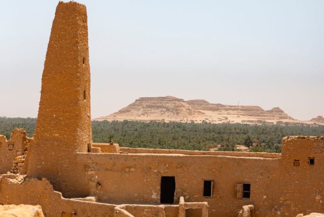 A distant view of a towering stone structure amidst the desert, with a green oasis of palm trees stretching out in the background.