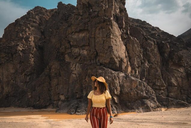 Woman wearing a sun hat and striped pants walking in a desert landscape with large rocky mountains in the background.