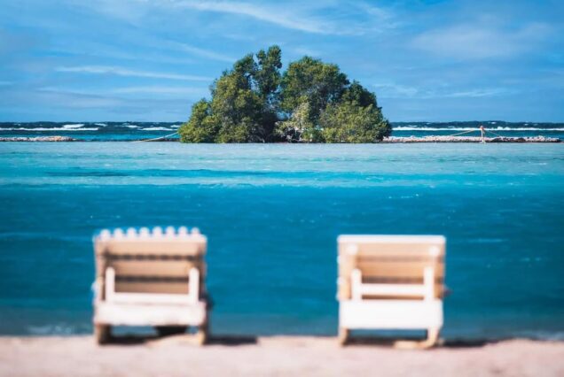 Two empty beach chairs facing a small island in turquoise blue water under a bright sky