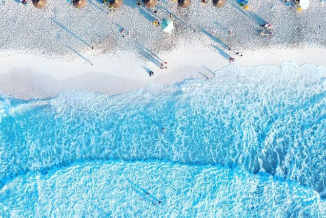 Aerial view of a vibrant blue ocean washing onto a white sandy beach with scattered sun umbrellas and people relaxing