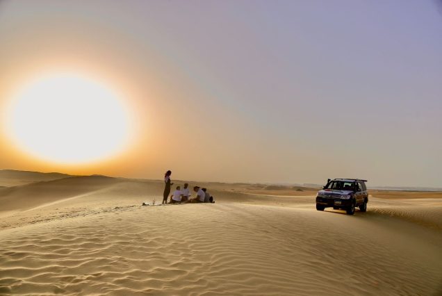 A group of people sitting on sand dunes at sunset, next to a 4x4 vehicle, enjoying the vast desert landscape with the sun setting low on the horizon.