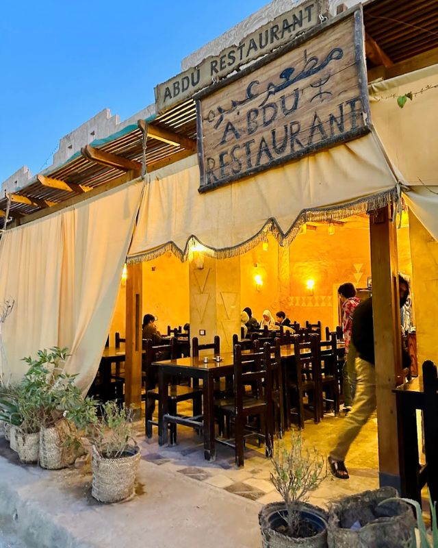 Entrance of Abdu Restaurant with warm yellow lighting, wooden tables, and chairs inside. Several patrons are dining in the open-air setting under a large canopy