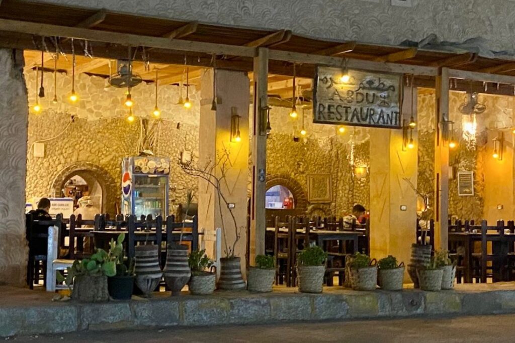 Exterior of Abdu Restaurant at night with soft, warm lighting and rustic stone walls visible inside. Wooden tables are set up for guests, with several plants in pots lining the entrance