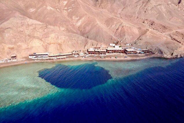 Aerial view of a beach with a deep blue coral reef offshore, nestled against desert mountains in Egypt