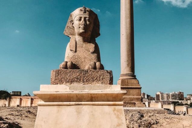 A stone sphinx statue with a tall column in the background, set against a clear blue sky