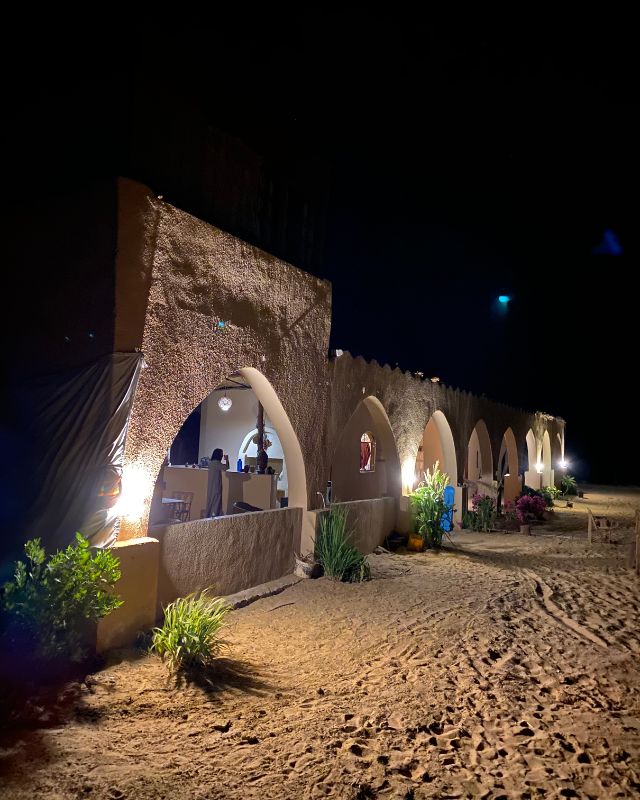 An arched building lit by soft lights at night, surrounded by desert sand, with plants and flowers growing near the entrance