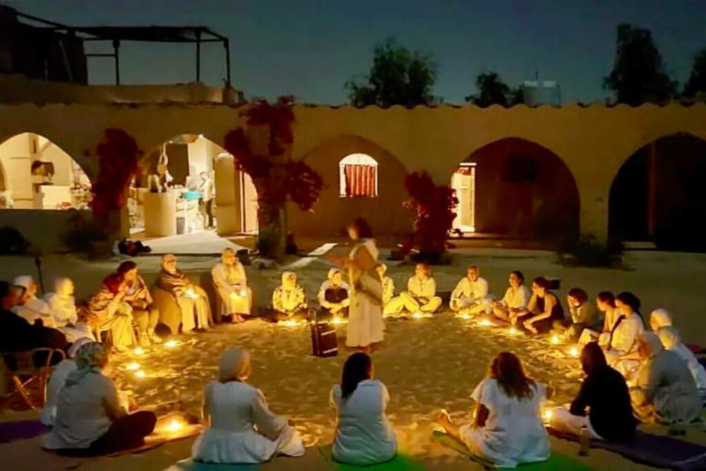 A group of people sitting in a large circle on the sand, surrounded by candlelight in an outdoor setting at night, participating in a gathering