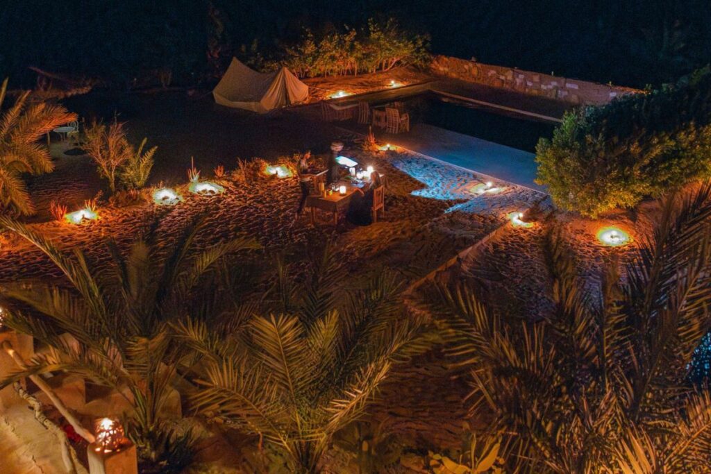 A romantic outdoor dinner setting lit by candles, surrounded by sand and palm trees at night. A single table is set for two, with a nearby tent and soft lighting creating a peaceful ambiance