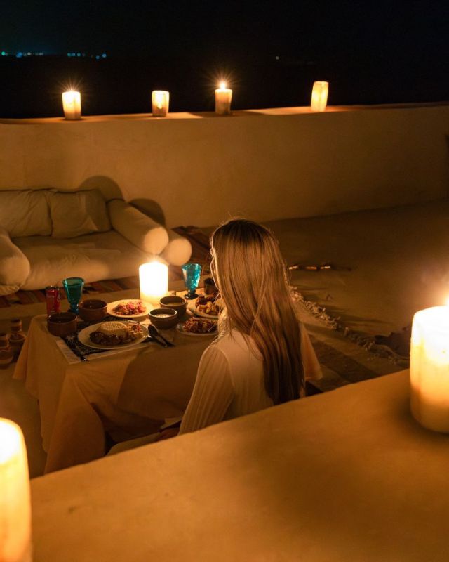 A woman seated at a low table for a candlelit dinner outdoors, with soft lighting and candles creating a peaceful ambiance under the night sky