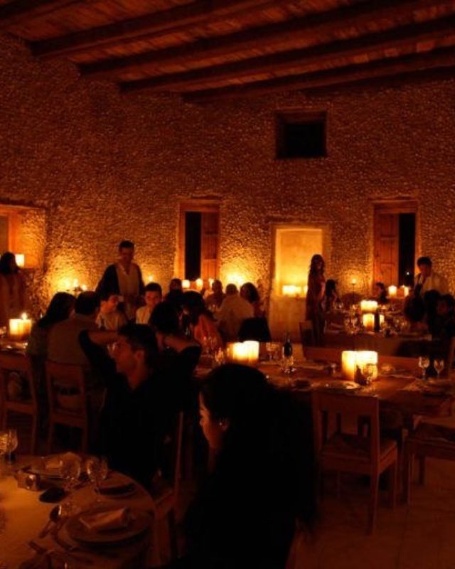A large group of diners gathered in a rustic, candlelit dining room with stone walls and wooden furniture, creating a warm and lively atmosphere