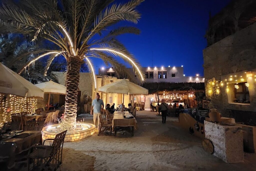 A cozy outdoor dining area under a beautifully lit palm tree at night, with warm lights strung around the restaurant, located in Siwa Oasis, Egypt