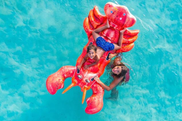 Two children floating on an inflatable red lobster in clear blue water, smiling and playing