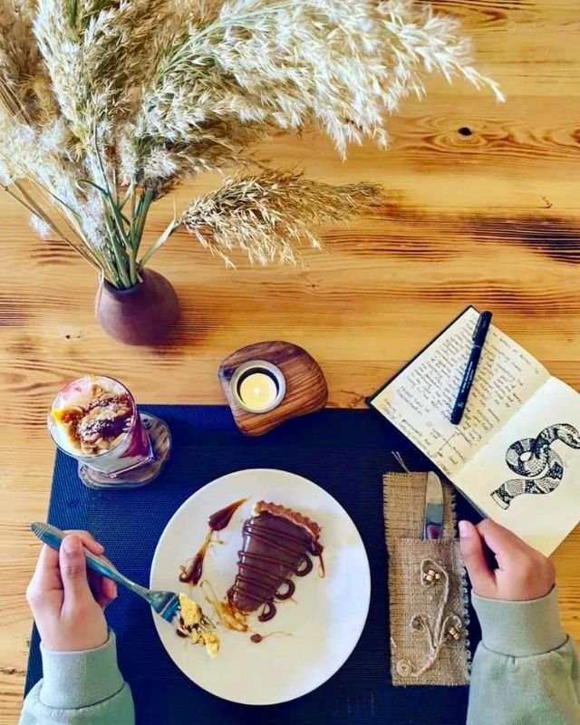 "Overhead shot of a dessert on a white plate, with a notebook and drink on a wooden table, creating a cozy ambiance