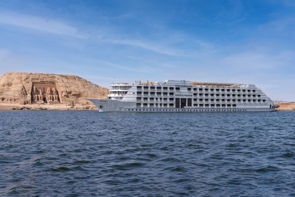 A large white cruise ship named "Steigenberger Omar El Khayam" sailing on the calm waters of Lake Nasser, with a rocky desert backdrop under a clear blue sky