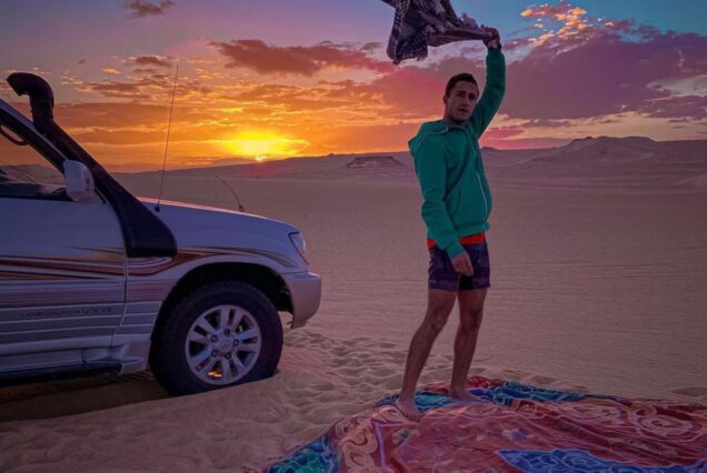 A man standing in front of a 4x4 in the desert, holding a scarf as the sun sets in the background