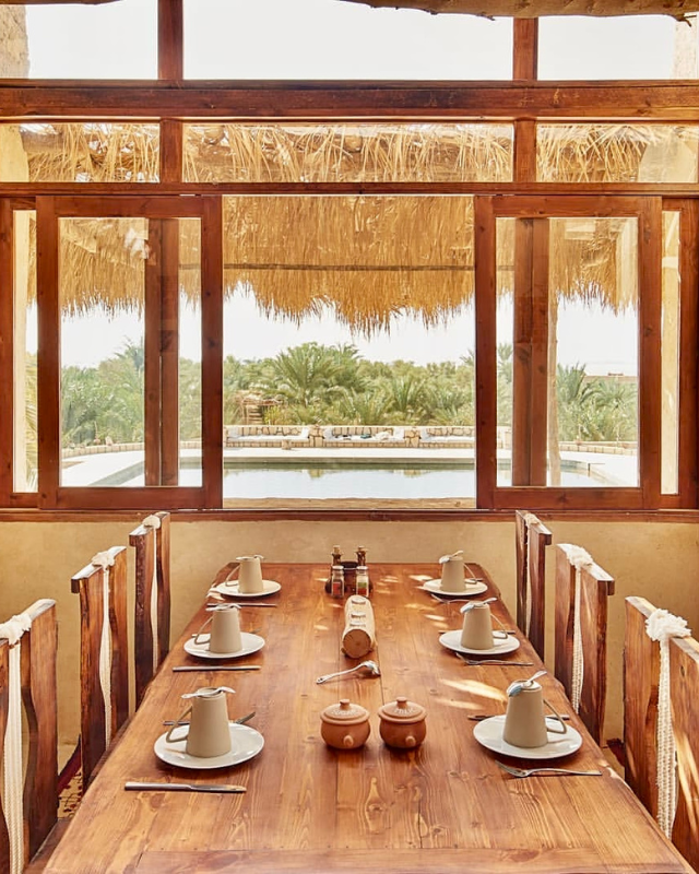 A wooden dining table set for eight, with a large window offering a view of a palm-fringed swimming pool and the desert landscape beyond