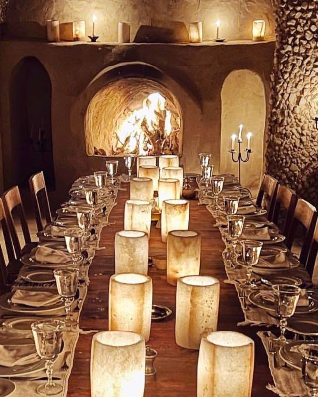 A beautifully set long dining table with candles, wine glasses, and place settings in front of a lit stone oven, creating a warm and intimate atmosphere