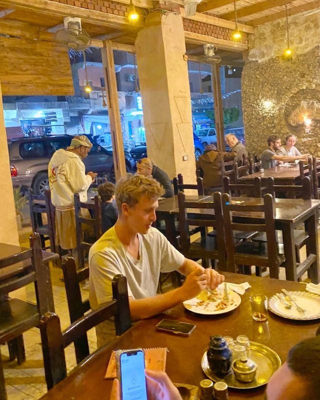 A male guest at Abdu Restaurant enjoying his meal at a wooden table, with other diners in the background. The cozy, rustic interior features stone walls and soft lighting