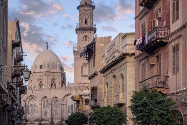 A street view of a historic mosque with a dome and a tall minaret, surrounded by old buildings at sunset