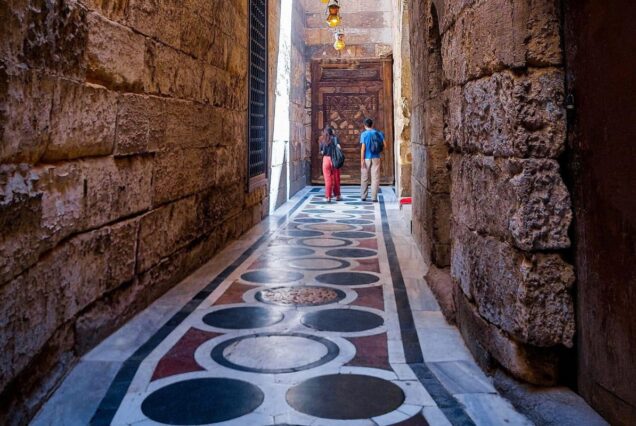 Narrow stone corridor with decorative tiled floor and two people walking towards a wooden door