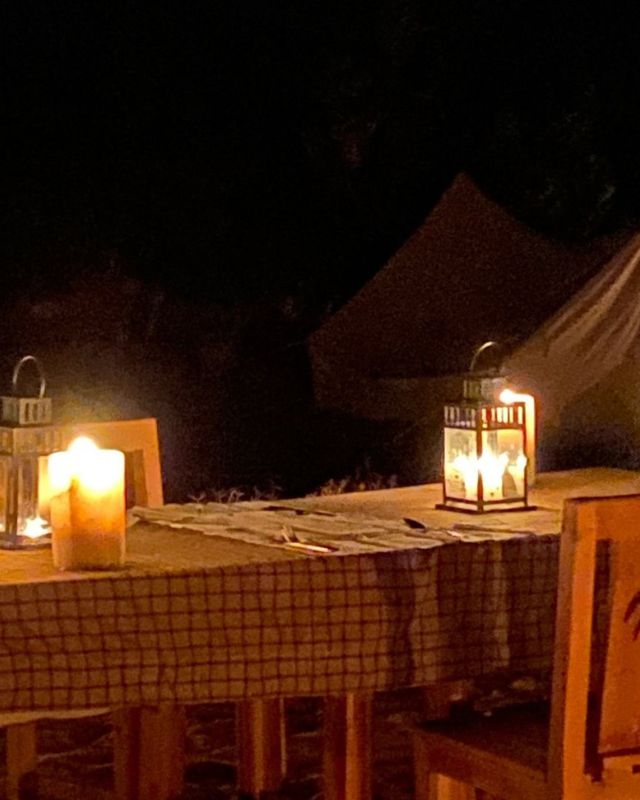 An outdoor dining table at night, lit by lanterns and candles, with a tent and trees in the background, creating a calm and serene desert atmosphere