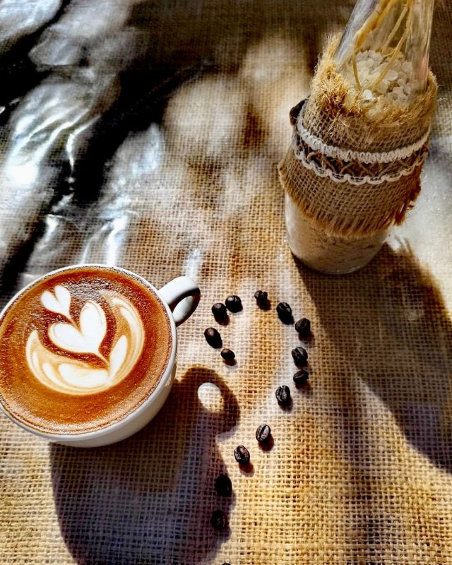 A cup of cappuccino with intricate latte art, placed on a burlap surface next to coffee beans arranged in a heart shape