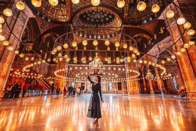 A grand hall illuminated with circular chandeliers in a mosque, with a woman standing in the center reaching upwards