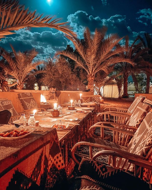 Nighttime dining setup under palm trees with candles illuminating the tables, creating a cozy and warm atmosphere under a moonlit sky