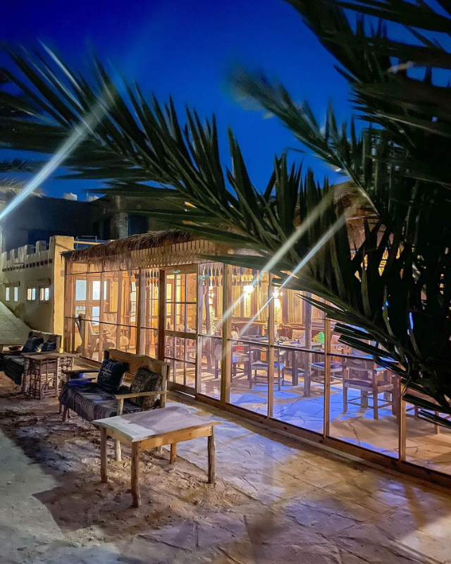 A dining area at night, illuminated by soft lighting, with palm trees framing a cozy outdoor seating space with wooden furniture and glass walls