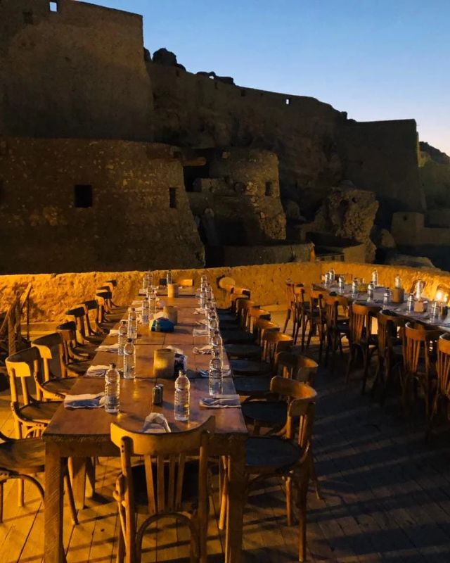 A large wooden dining table set for an outdoor meal against the backdrop of ancient stone ruins at dusk