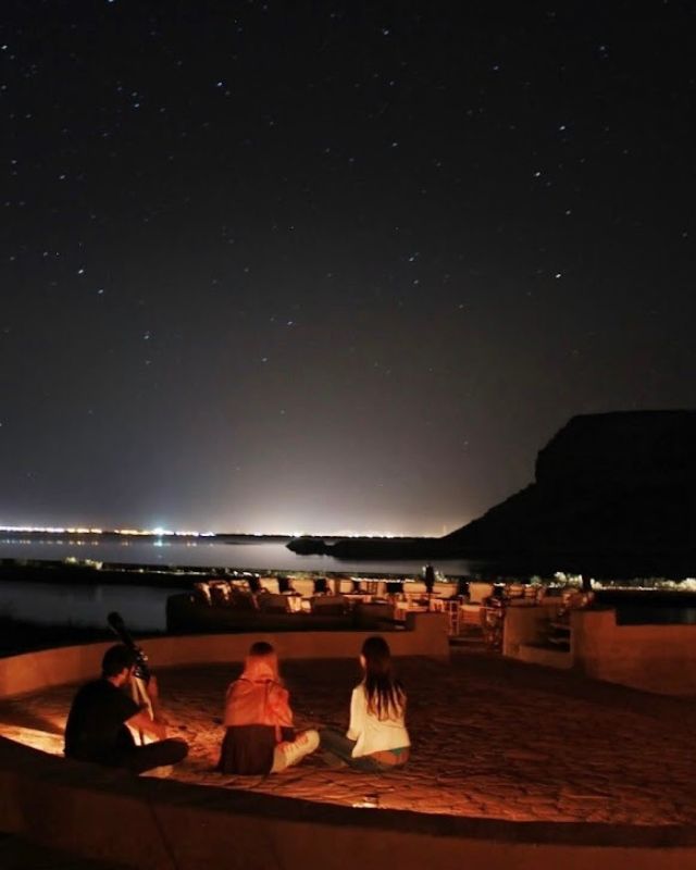 A starry night sky above a group of people seated around a fire near the water