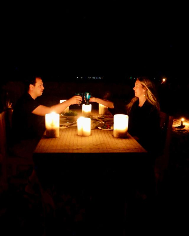 A couple enjoying a candlelit dinner in the desert at night, toasting with glasses over a table illuminated by warm candlelight