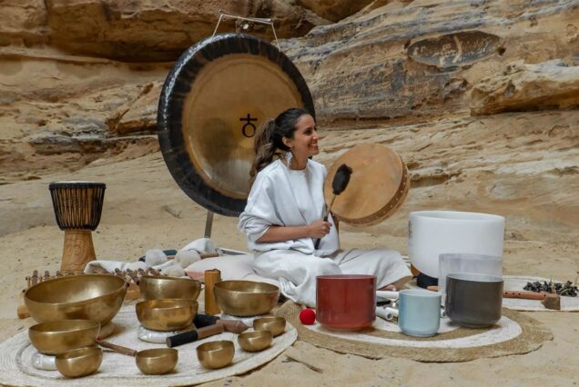 A woman seated with a collection of musical instruments, including Tibetan singing bowls, a drum, and a gong, in a desert canyon