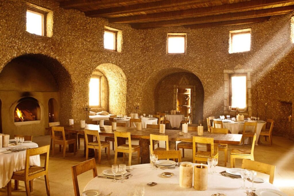 A sunlit dining room with rustic stone walls, wooden chairs, and tables, featuring a lit fireplace and beams of sunlight streaming through the windows