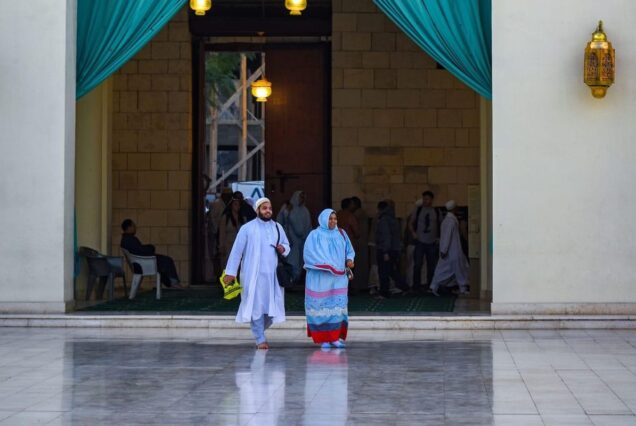 Man and woman in traditional attire walking outside a mosque entrance with turquoise drapes