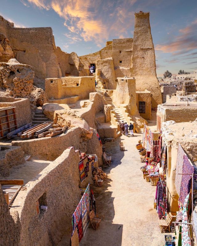 An ancient, rustic village built from stone, with narrow pathways and colorful textiles displayed along the walls