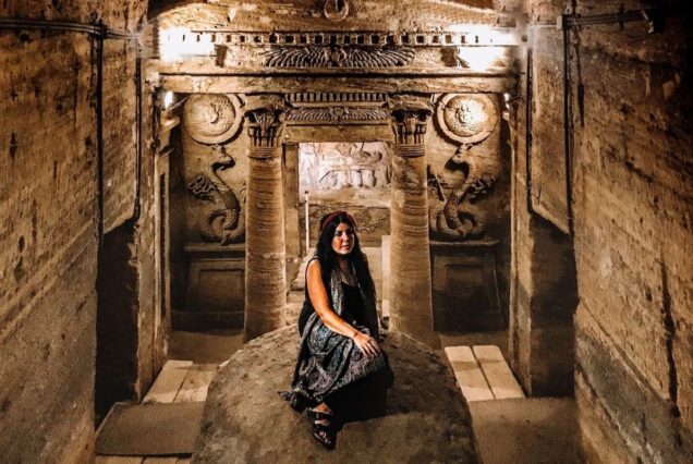 A woman sitting on a stone inside an ancient underground chamber adorned with carvings and pillars