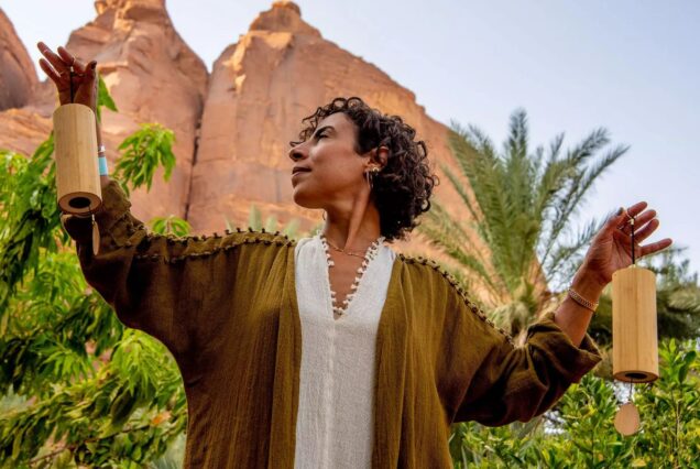 A woman in a flowing robe holding chimes with a serene expression, set against a backdrop of rocky cliffs and palm trees